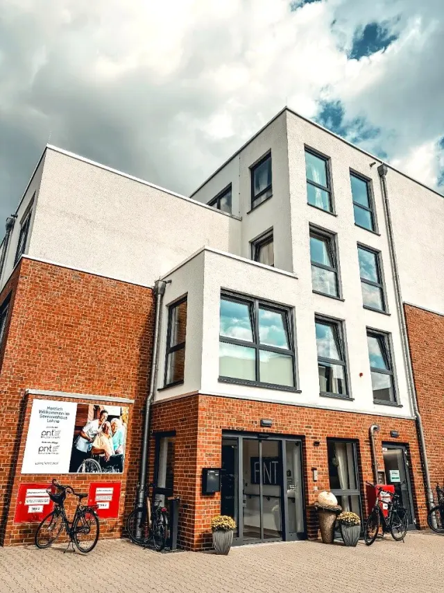 Freundliche Pflegefachkraft vor dem Haupteingang und der Außenfassade des Seniorenhauses